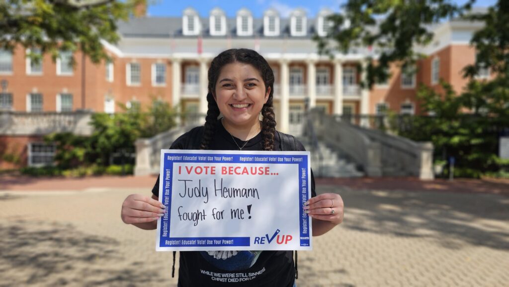 A young woman holds a sign with the words "I vote because Judy Heumann fought for me!"