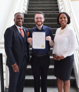 A white man holds an official-looking document in a frame. An African American man stands on his right, and an African American woman stands on his left.