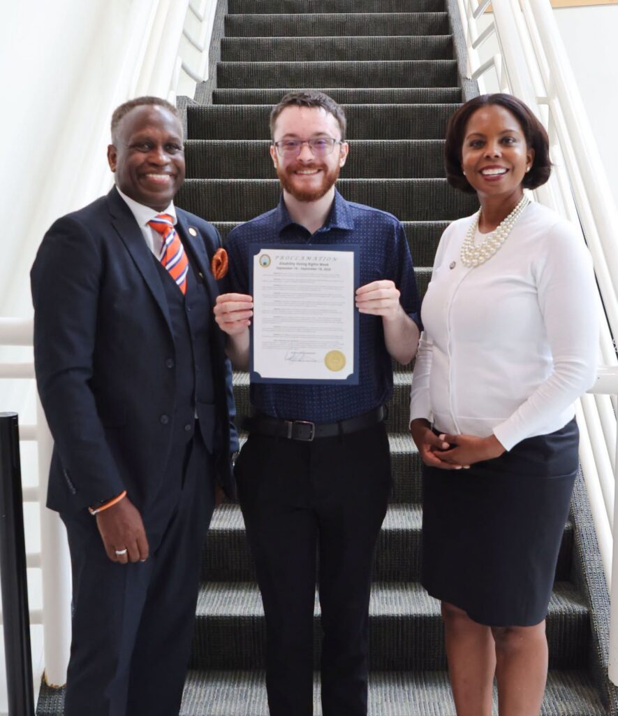 A white man holds an official-looking document framed in blue cardboard. An African American man stands on his right, and an African American woman stands on his left.